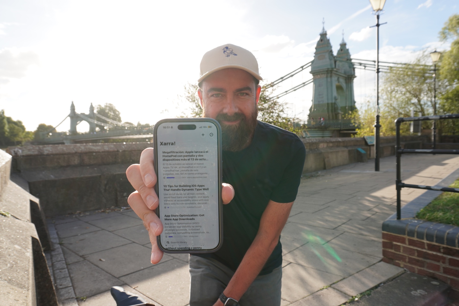 Dani smiling while holding an iPhone showing Xarra, with Hammersmith Bridge behind him.