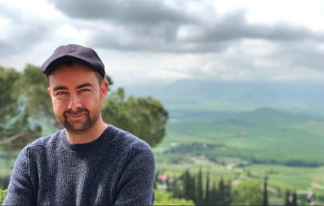 Daniel, smiling slightly while wearing a navy sweater and a flat cap, standing outdoors with the rolling green hills of Tuscany and a cloudy sky in the background.